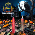 Two Halloween taper candles, one unlit red and one burning with red wax drips, displayed against a moonlit backdrop with pumpkins and an owl.