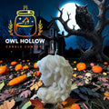 An Bone white, textured wax candle shaped like a stylized face is displayed in a spooky Halloween scene with pumpkins, autumn leaves, a haunted house, and a large owl on a tree branch under a full moon.