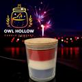 Red, white, and blue layered candle in a clear glass container with a wooden lid, displayed against fireworks with Owl Hollow Candle Company branding.