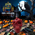 A red skull-shaped candle with a coiled snake wrapped through it, set in a Halloween-themed scene with pumpkins, autumn leaves, a haunted house, an owl on a tree branch, and a full moon.