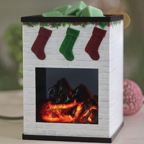 White fireplace-shaped wax warmer with glowing logs, red and green Christmas stockings, and green wax melts in the top tray.